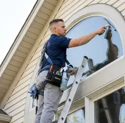 Professional window cleaner on a ladder using a squeegee to clean an exterior arched window, Albuquerque, NM.
